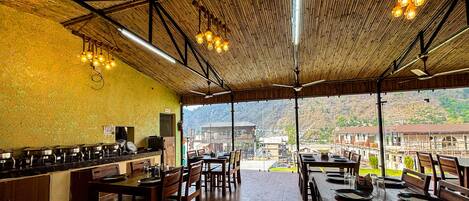 A restaurant with a bamboo ceiling, wooden tables and chairs, and a view of buildings and hills in the background.