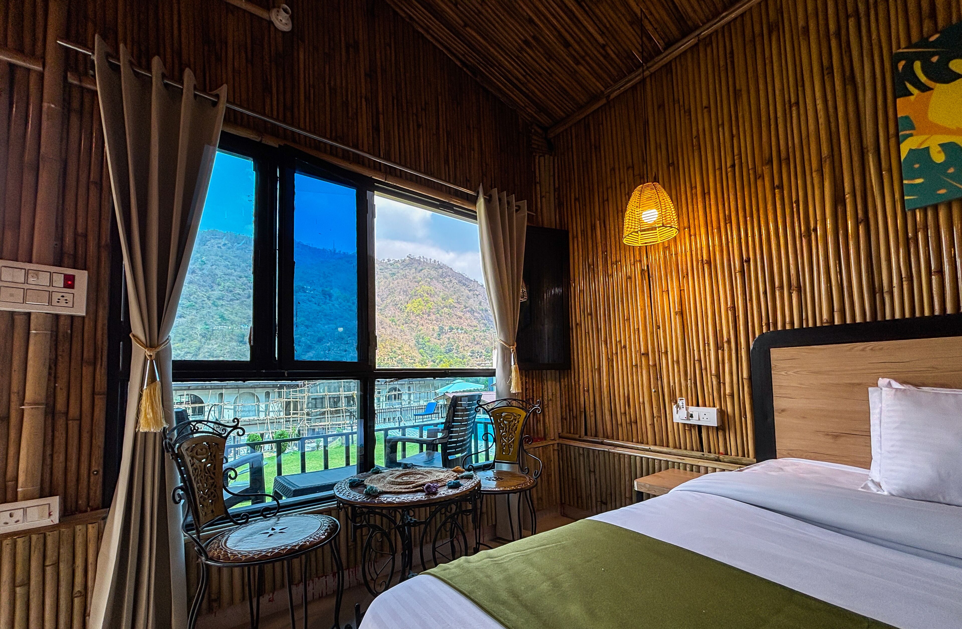 A room with a bed, a table with chairs, a window with curtains, a wall-mounted light, and a view of a mountainous landscape.