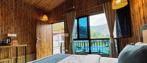 A wooden room with a bed, a window with a view of a pool and mountains, and a hanging light fixture.