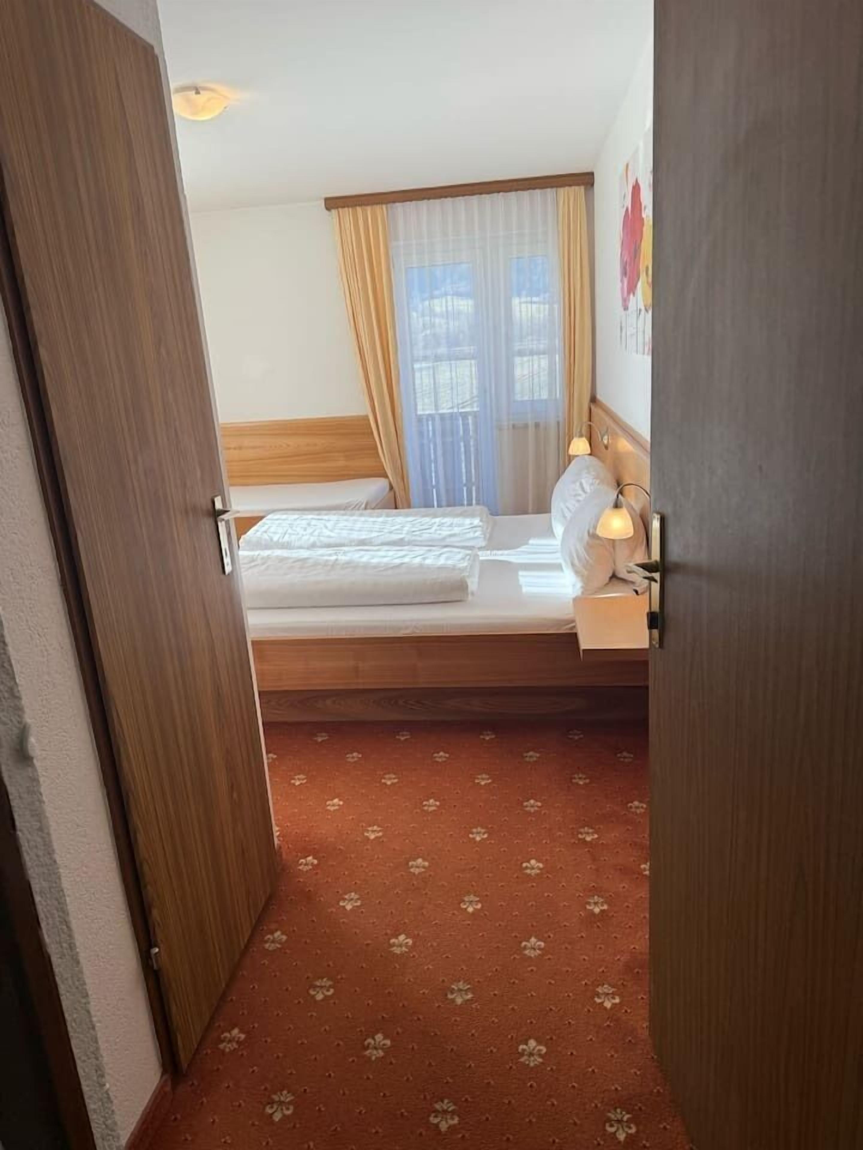 Ein Hotelzimmer mit Bett, einem Fenster mit Vorhängen, einer Wandlampe und einem gemusterten Teppich.