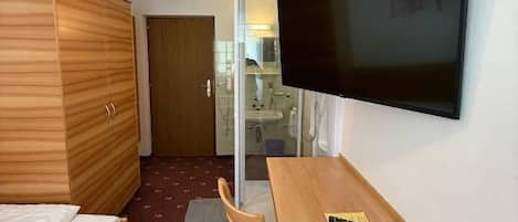 A compact hotel room with a bed, a desk, a chair, a television, and a bathroom visible through an open door.