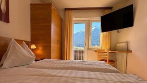 Une chambre d’hôtel avec un lit, un téléviseur fixé au mur, une fenêtre donnant sur les montagnes et une armoire en bois.