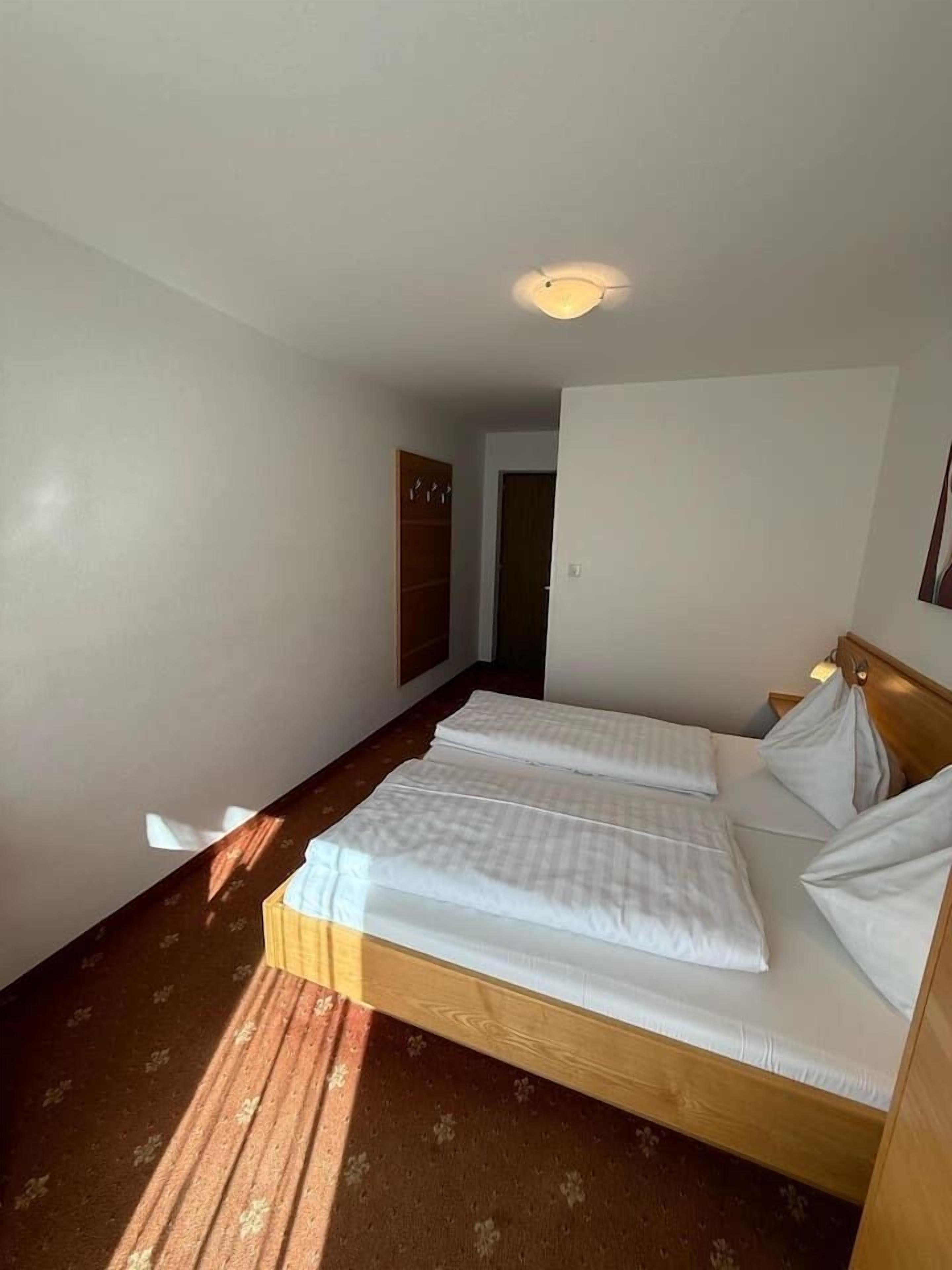 A hotel room with a double bed, a wooden wardrobe, and a small desk with a lamp.