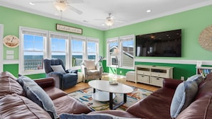A living room with a green wall, white ceiling, and large windows. There is a brown leather sofa, a white coffee table, and a blue armchair. - Arches: Oceanfront retreat in Oak Island, 4BR, 3BA, sleeps 12, ideal for families. (Oak Island)