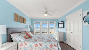 A bedroom with a bed, a ceiling fan, a dresser, a nightstand, and a view of the ocean through large windows. - Arches: Oceanfront retreat in Oak Island, 4BR, 3BA, sleeps 12, ideal for families. (Oak Island)