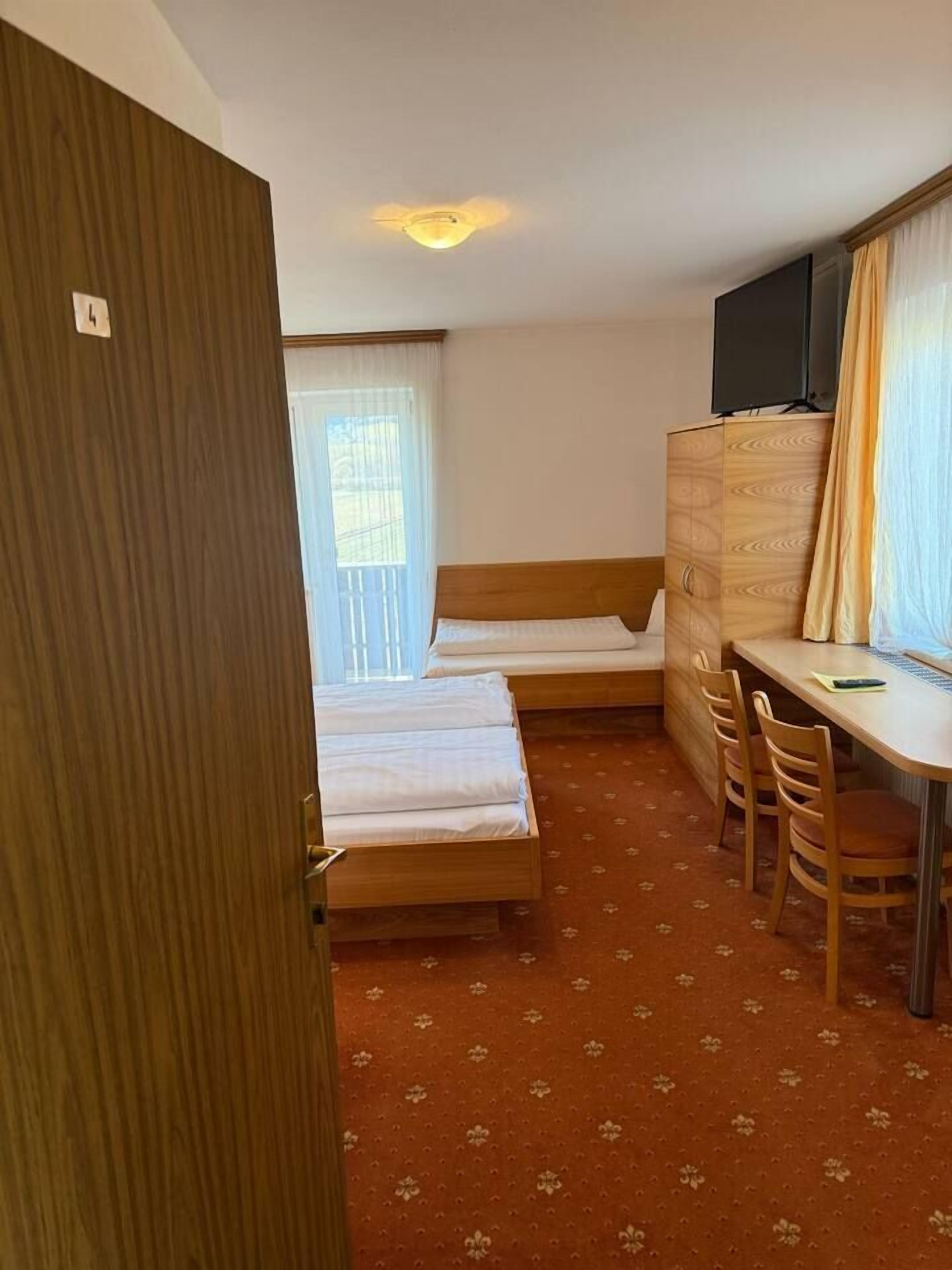 A hotel room with two single beds, a wooden desk with a chair, a TV on a cabinet, and a window with curtains.