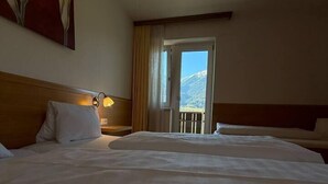 A hotel room with a large bed, a bedside table, a lamp, a window with a view of mountains, and a door leading to a balcony.
