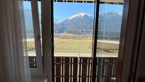 A room with a sliding glass door leading to a balcony with a view of a mountain and a field.