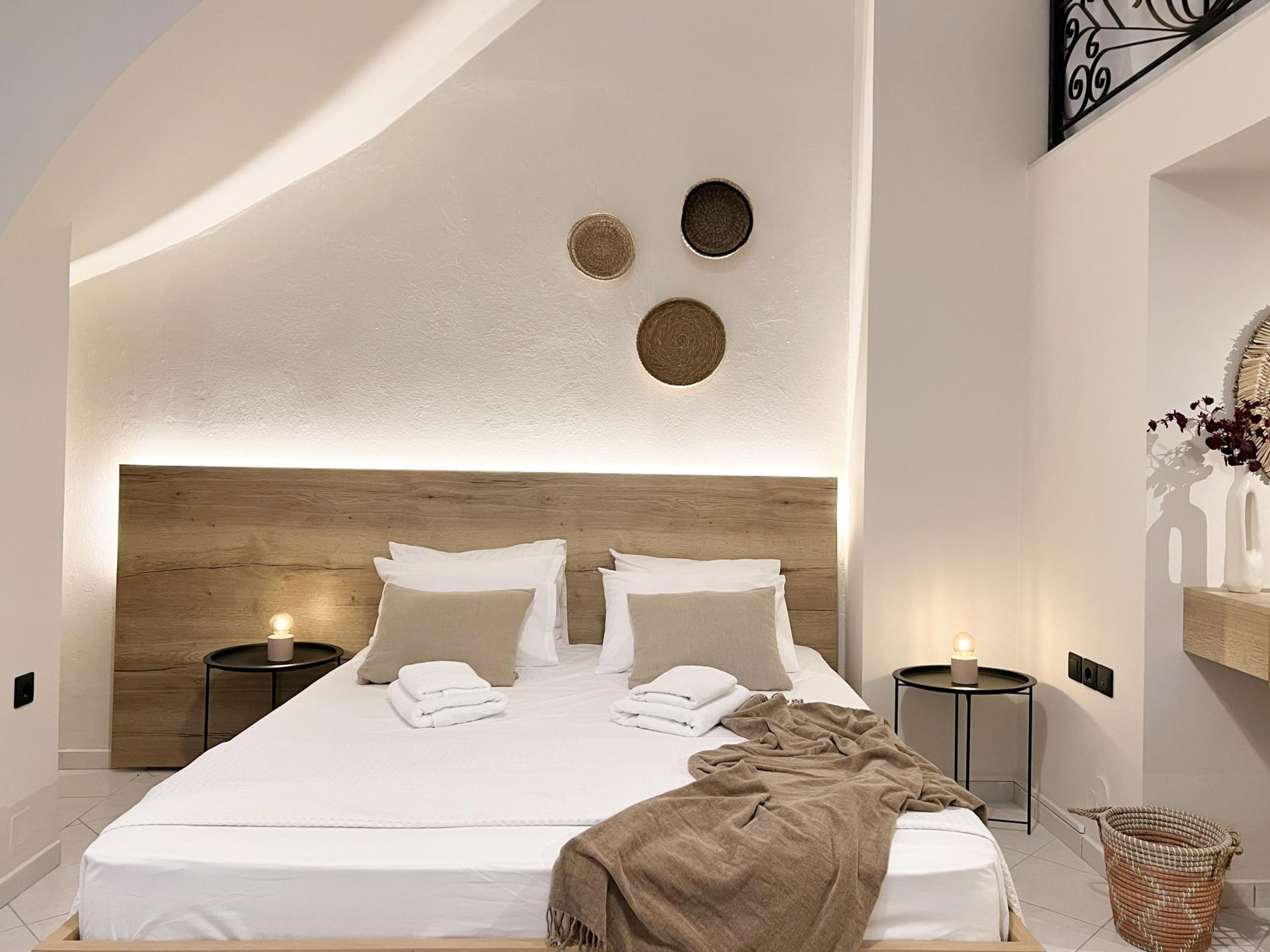 Une chambre à coucher moderne avec une tête de lit en bois, deux tables de chevet avec des lampes, un lit recouvert de serviettes blanches soigneusement pliées et un panier en osier posé sur le sol.