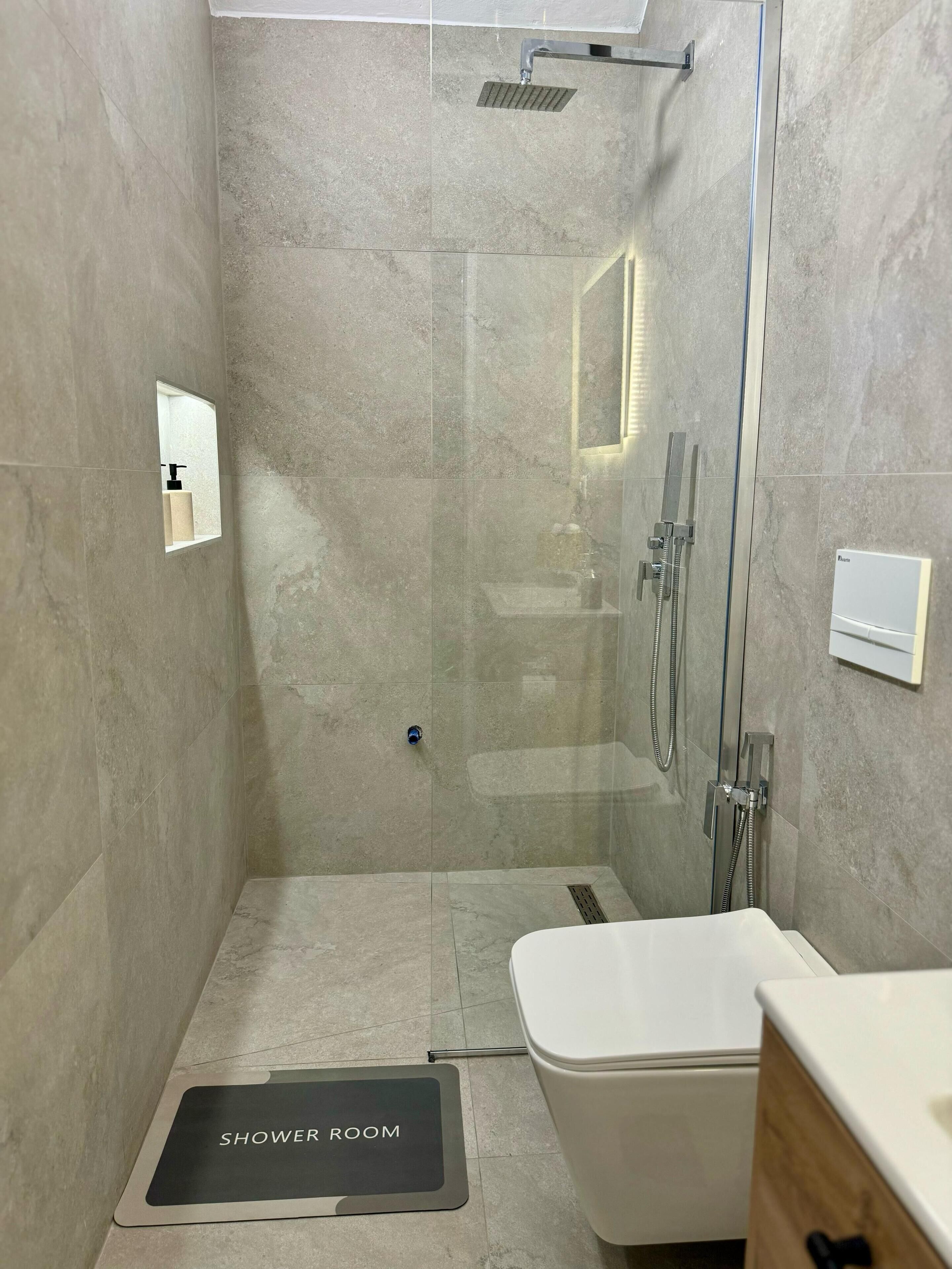A modern shower room with a glass door, a handheld showerhead, and a handheld showerhead on the wall. There is a small mirror on the wall and a towel rack.