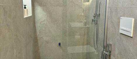 A modern shower room with a glass door, a handheld showerhead, and a handheld showerhead on the wall. There is a small mirror on the wall and a towel rack.
