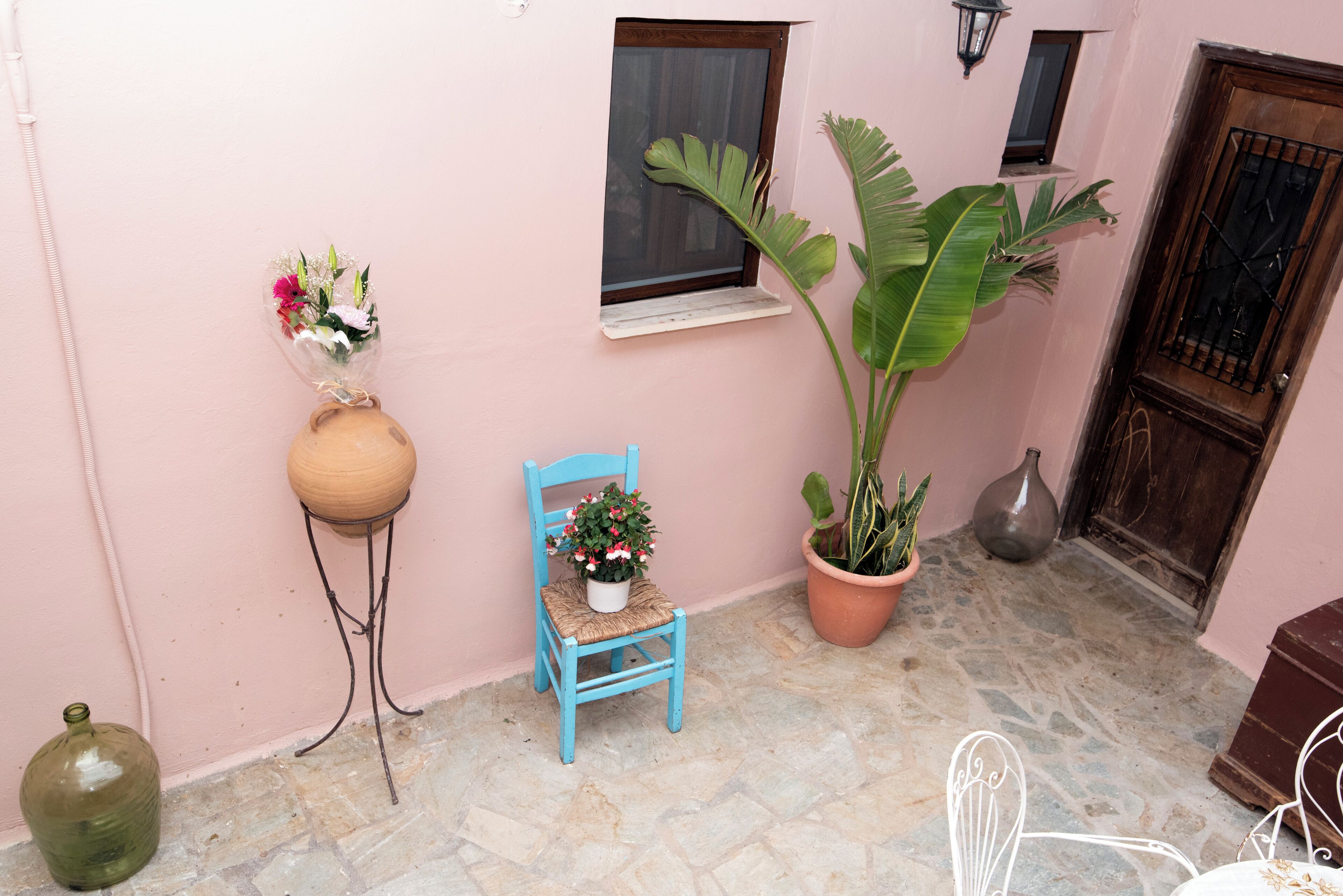 Une pièce aux murs roses, avec une petite fenêtre, une chaise sur laquelle est posée une plante en pot, une grande plante en pot et un vase rempli de fleurs.