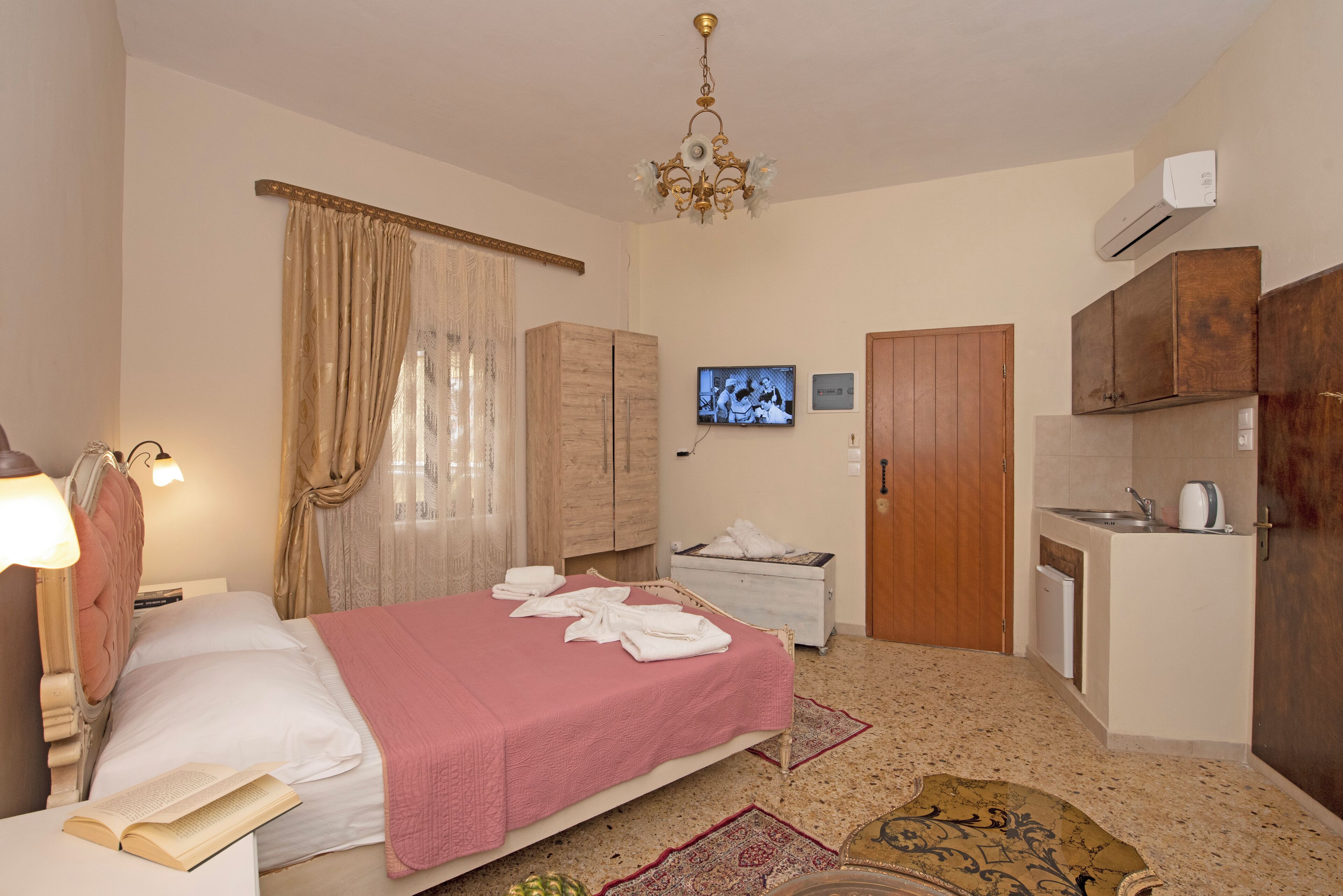 A hotel room with a bed, a bedside table with a book, a TV, a wardrobe, a small kitchenette, and a bathroom.