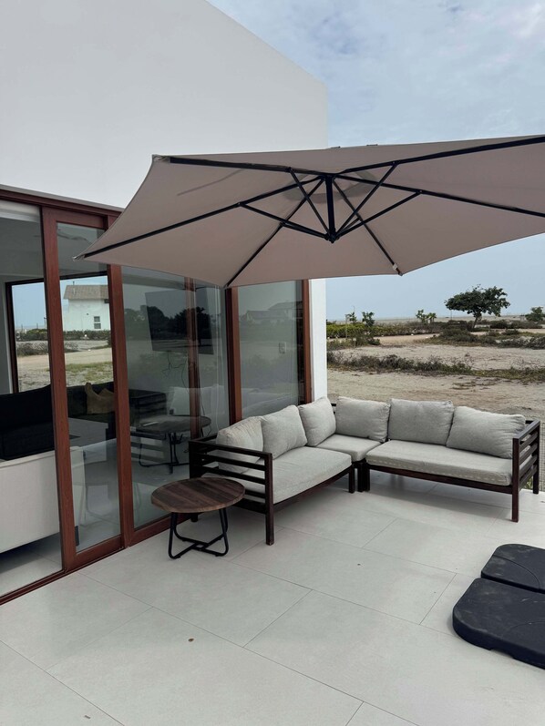 A modern outdoor seating area with a sofa and a table, a large umbrella, and a view of the beach.