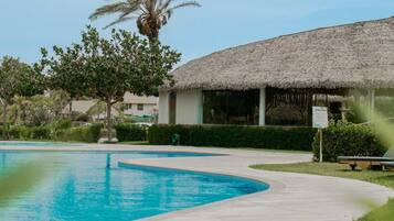Una piscina amb vora corbada, un edifici amb teulada de palla i una palmera.