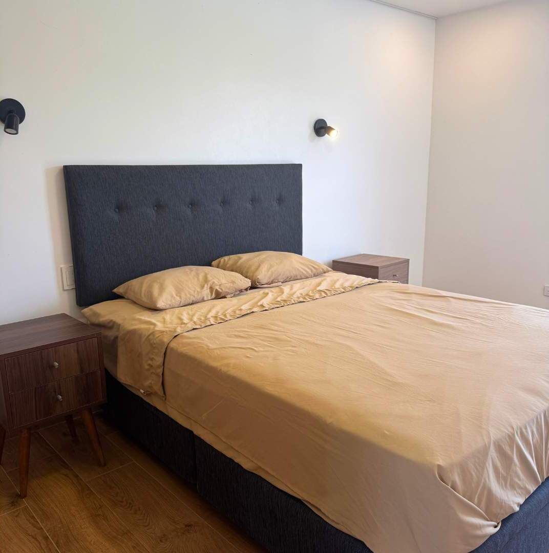 Un letto rifatto con cura, una testiera scura, un comodino in legno e un punto luce a parete.