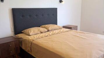 Un letto rifatto con cura, una testiera scura, un comodino in legno e un punto luce a parete.