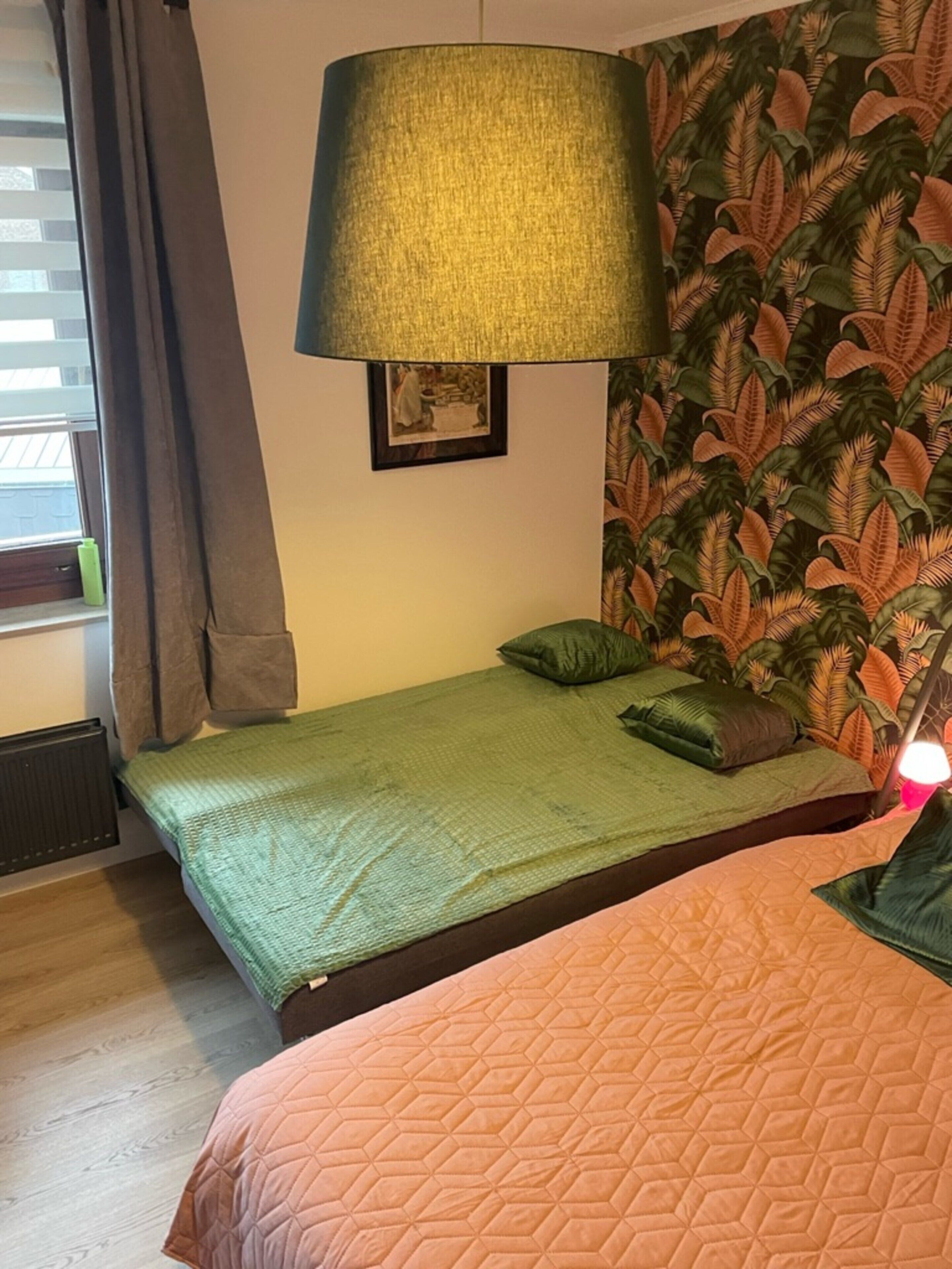Une chambre avec deux lits, une couvre-lit verte, une couvre-lit marron, une grande lampe fixée au mur et un papier peint orné de motifs de feuilles tropicales.