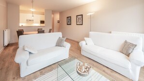 A modern living room with two white sofas, a glass coffee table, and a kitchen in the background.