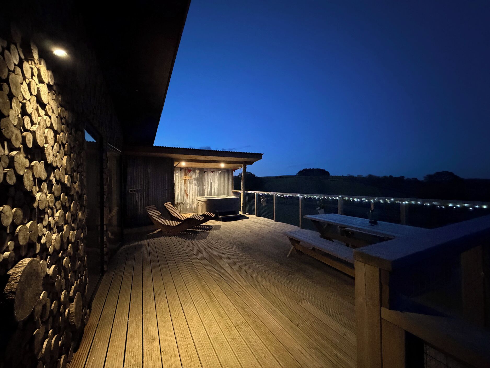 A wooden deck with a hot tub and lounge chairs, surrounded by stone walls and string lights.