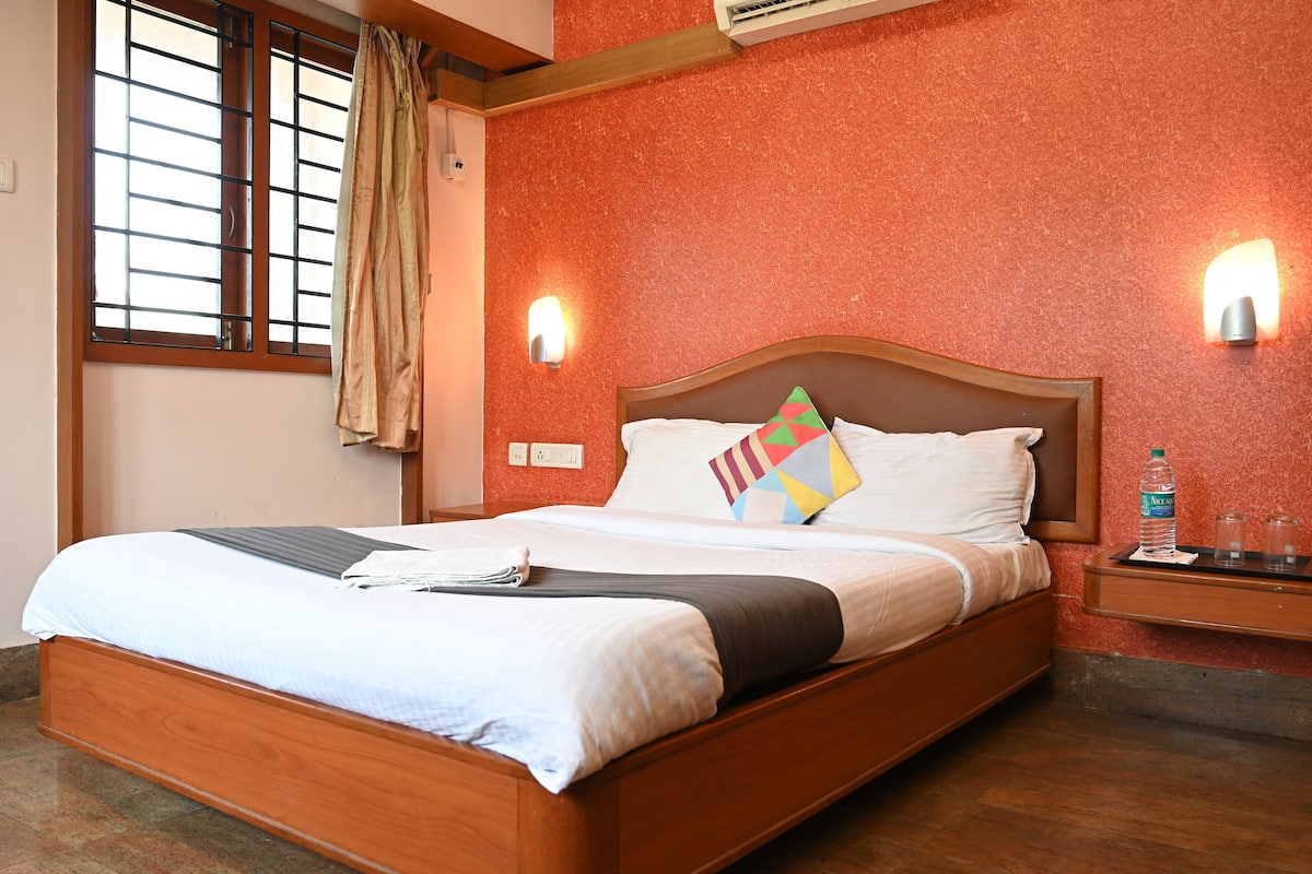 A neatly made bed with a wooden frame, two wall-mounted lights, a window with a curtain, and a small bedside table with a water bottle and glasses.