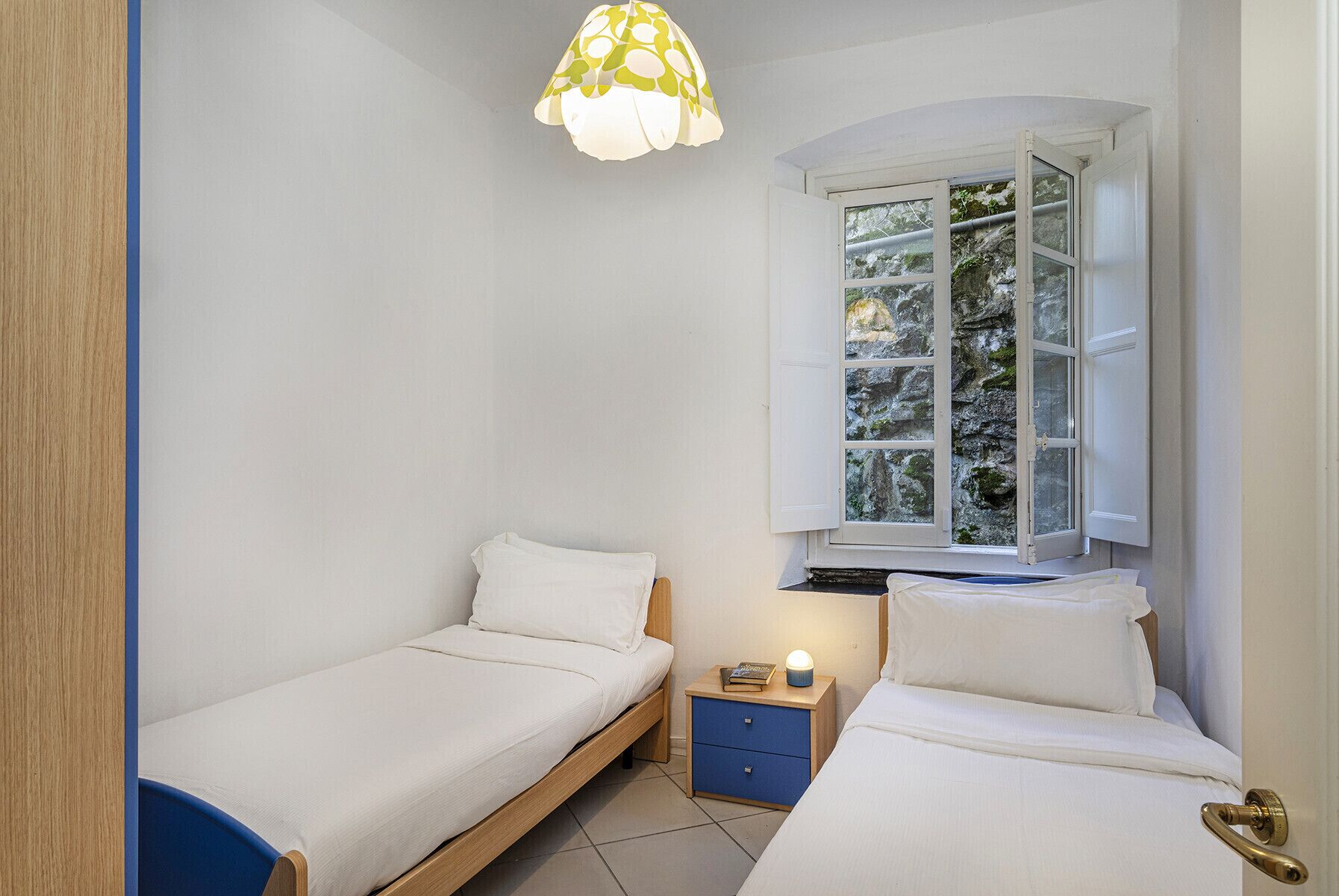 Een kleine, goed verlichte slaapkamer met twee eenpersoonsbedden, een raam met uitzicht op groen en een nachtkastje met een lamp.