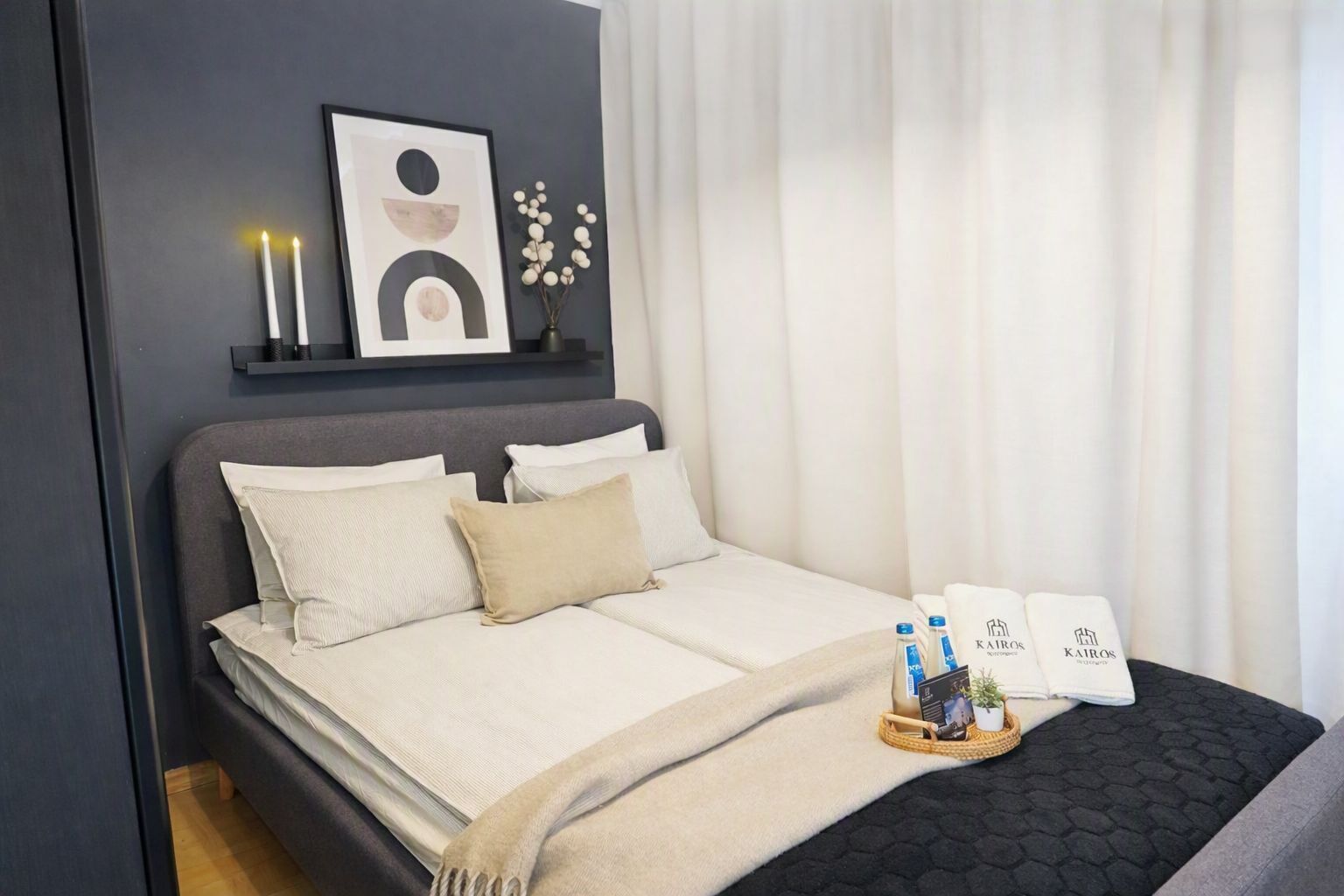 A modern hotel room with a bed, pillows, a bedside table with candles, a framed artwork, and a tray with toiletries.