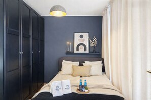 Habitación de hotel con cama, armario oscuro, obras de arte en la pared, velas y una bandeja con toallas y botellas.