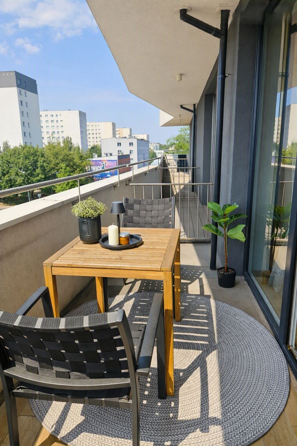 A balcony with a wooden table, two chairs, a potted plant, and a small table with a candle and a container.