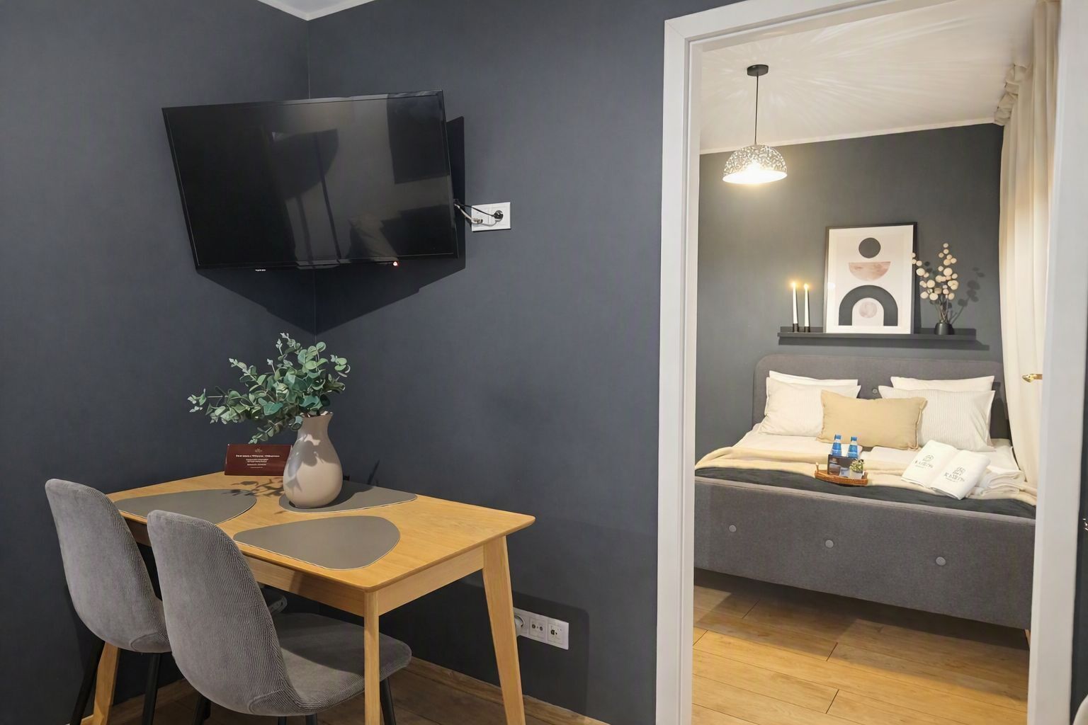 A room with a bed, a TV mounted on the wall, a table with chairs, and a potted plant.