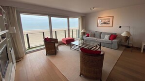 A living room with a sofa, a coffee table, a wicker ottoman, a floor lamp, and a view of the beach. - Seafront Palm Beach Stay (Knokke-Heist)