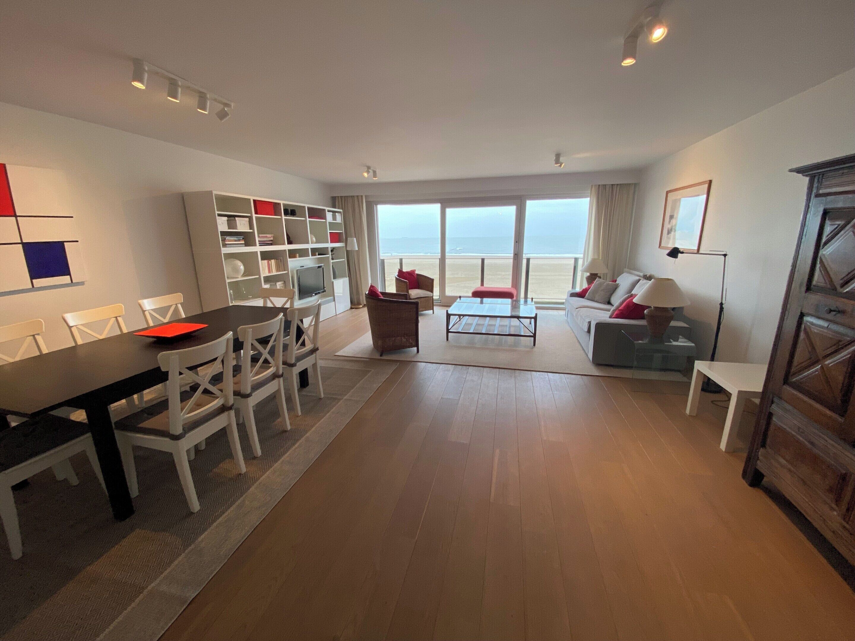 A spacious living area with a dining table, chairs, a sofa, and a TV. There is a large window with a view of the beach.