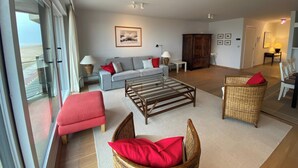 A spacious living room with a grey sofa, red cushions, a wooden coffee table, wicker chairs, and a view through large windows. - Seafront Palm Beach Stay (Knokke-Heist)