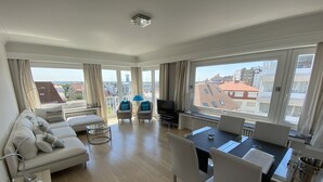 A modern living room with a sofa, two armchairs, a dining table with chairs, a TV, and large windows offering a city view. - Apartment in Knokke Near the Beach (Knokke-Heist)
