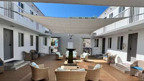 Un espacio al aire libre techado con mobiliario de mimbre y una mesa central, rodeado de paredes blancas y balcones.