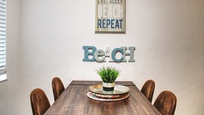 Un comedor con una mesa de madera, cuatro sillas, una planta en maceta y una pared con un cartel que dice 'BEACH' y otro que dice 'EAT, SLEEP, BEACH, REPEAT'.