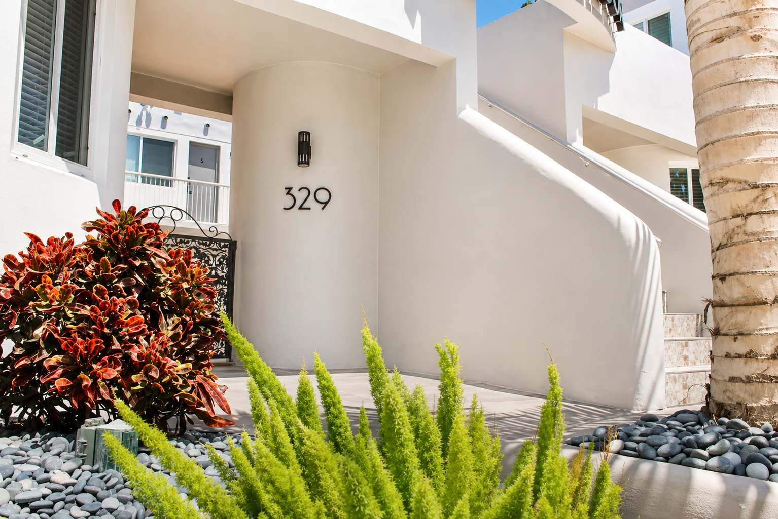 A modern building with a large number '329' on the wall, a staircase, and a well-maintained garden with red-leafed shrubs and green plants.