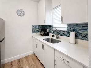 Una cocina con un lavamanos de acero inoxidable, armarios blancos, una cafetera y un reloj en la pared.