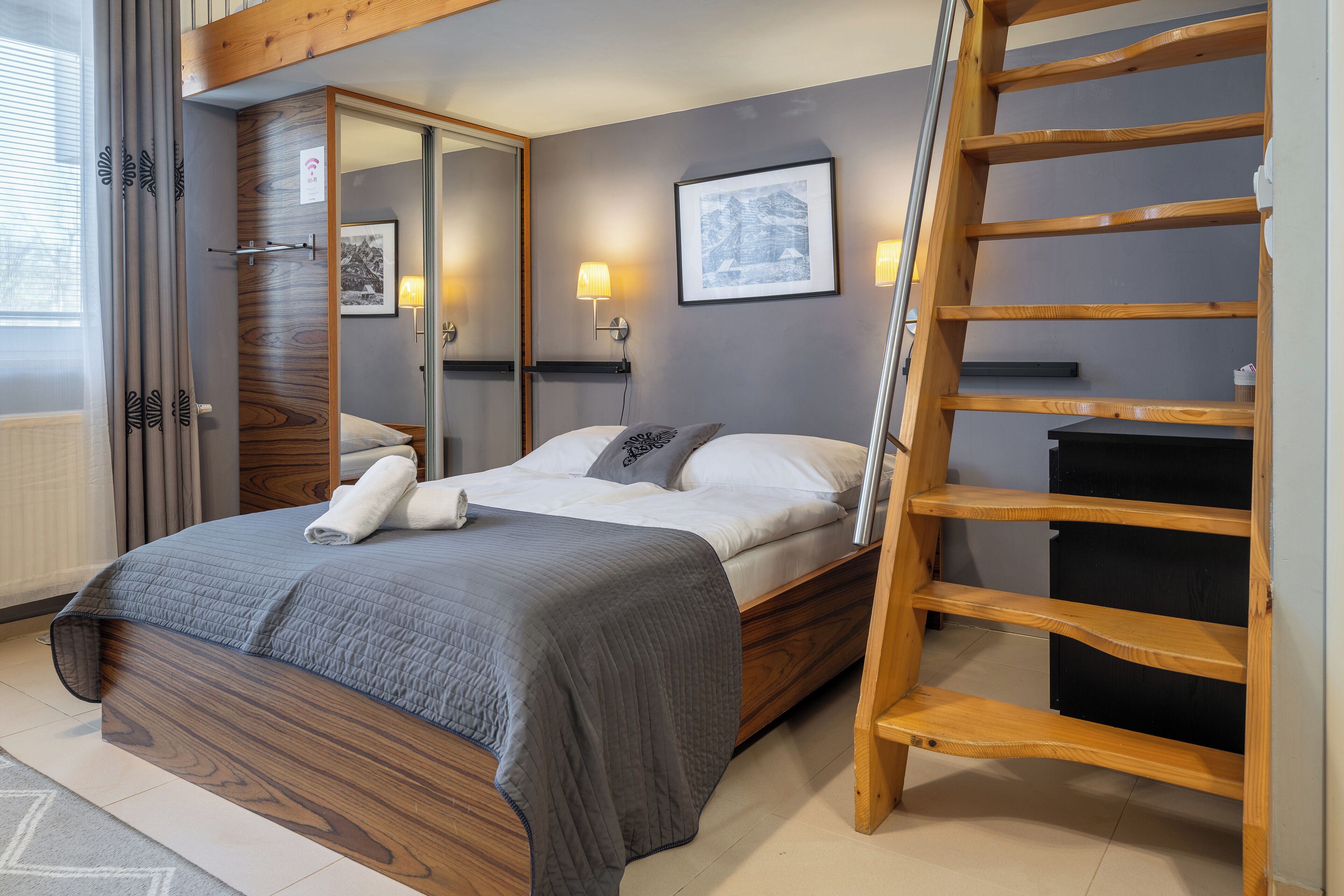 A bedroom with a wooden bed, a ladder, a mirror, and a framed picture on the wall.