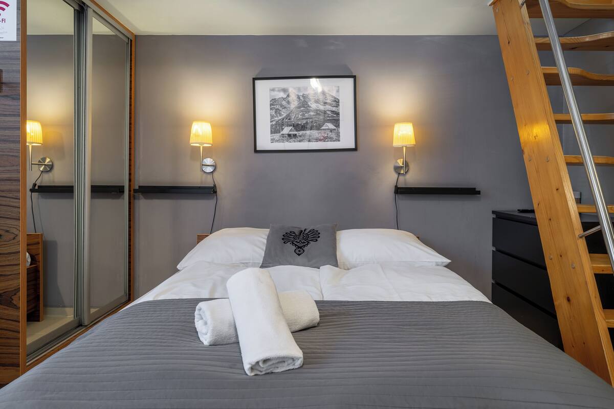Una cama bien tendida con una toalla doblada, un cuadro en la pared y dos lámparas adosadas a la pared.