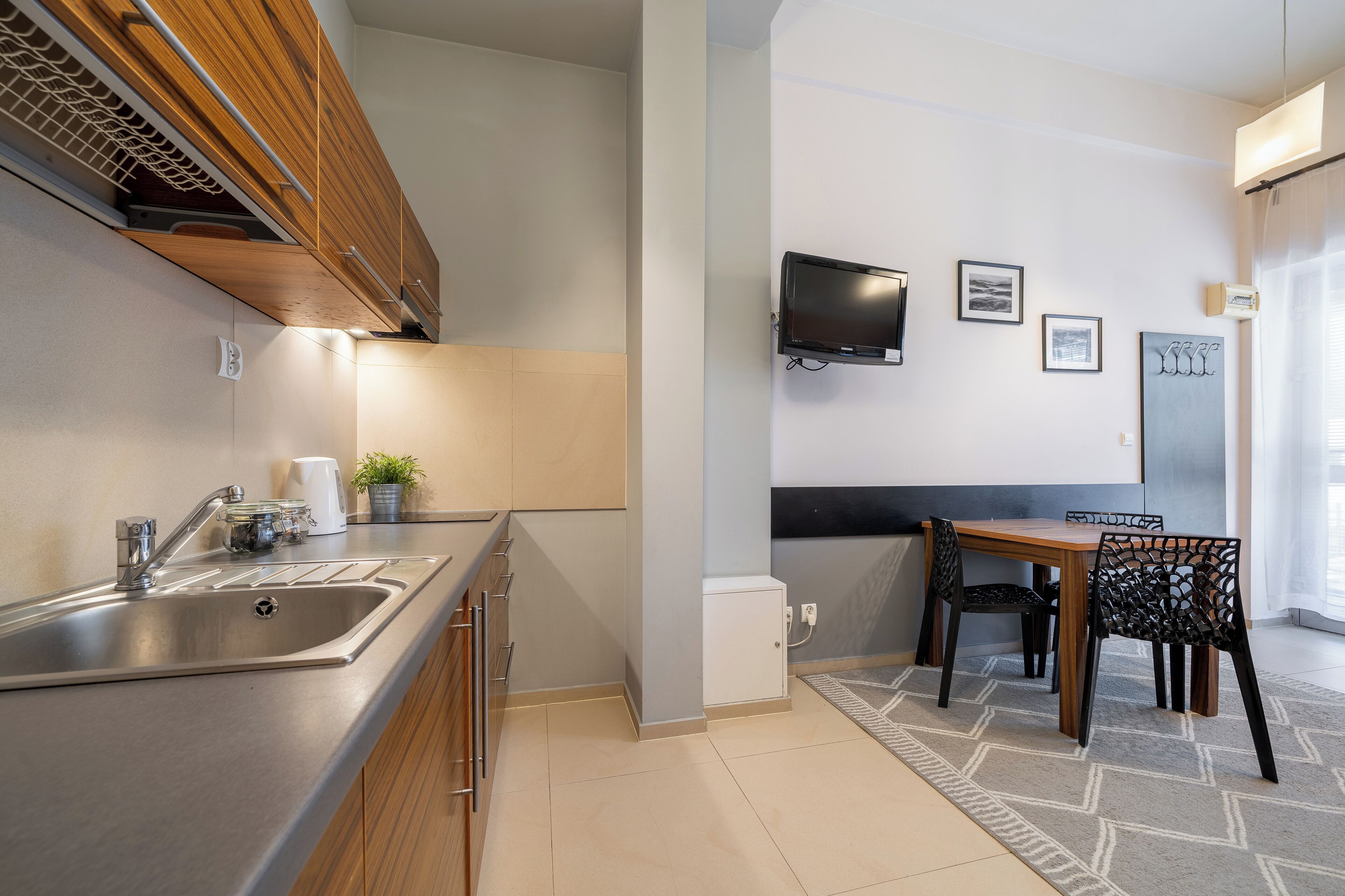 Une cuisine moderne dotée d’un évier en acier inoxydable, de meubles en bois et d’un petit réfrigérateur blanc. On y trouve une table à manger avec des chaises, ainsi qu’un téléviseur à écran plat fixé au mur.