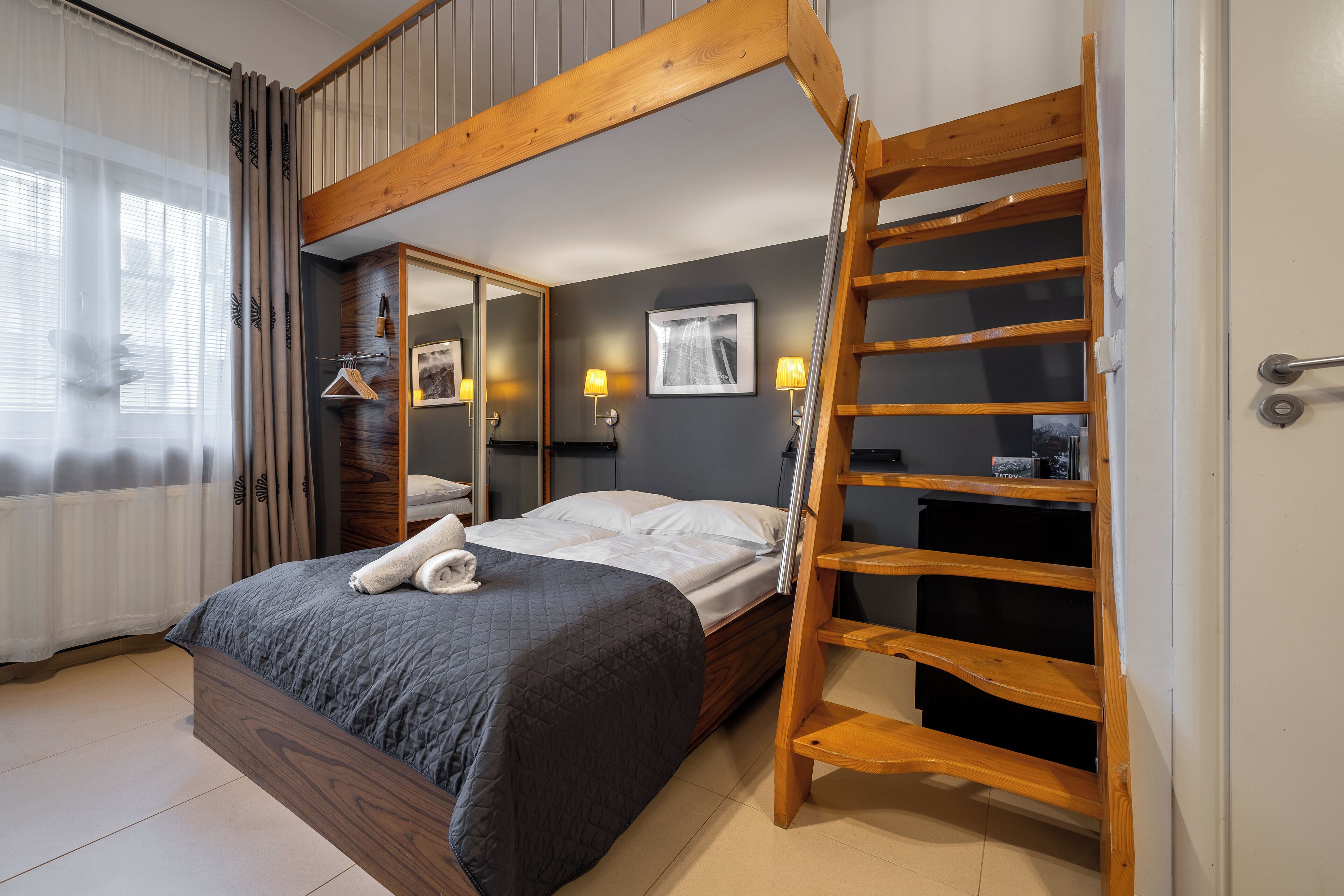 A bedroom with a wooden bunk bed, a ladder, a bedside table, a mirror, and a wall-mounted shelf.
