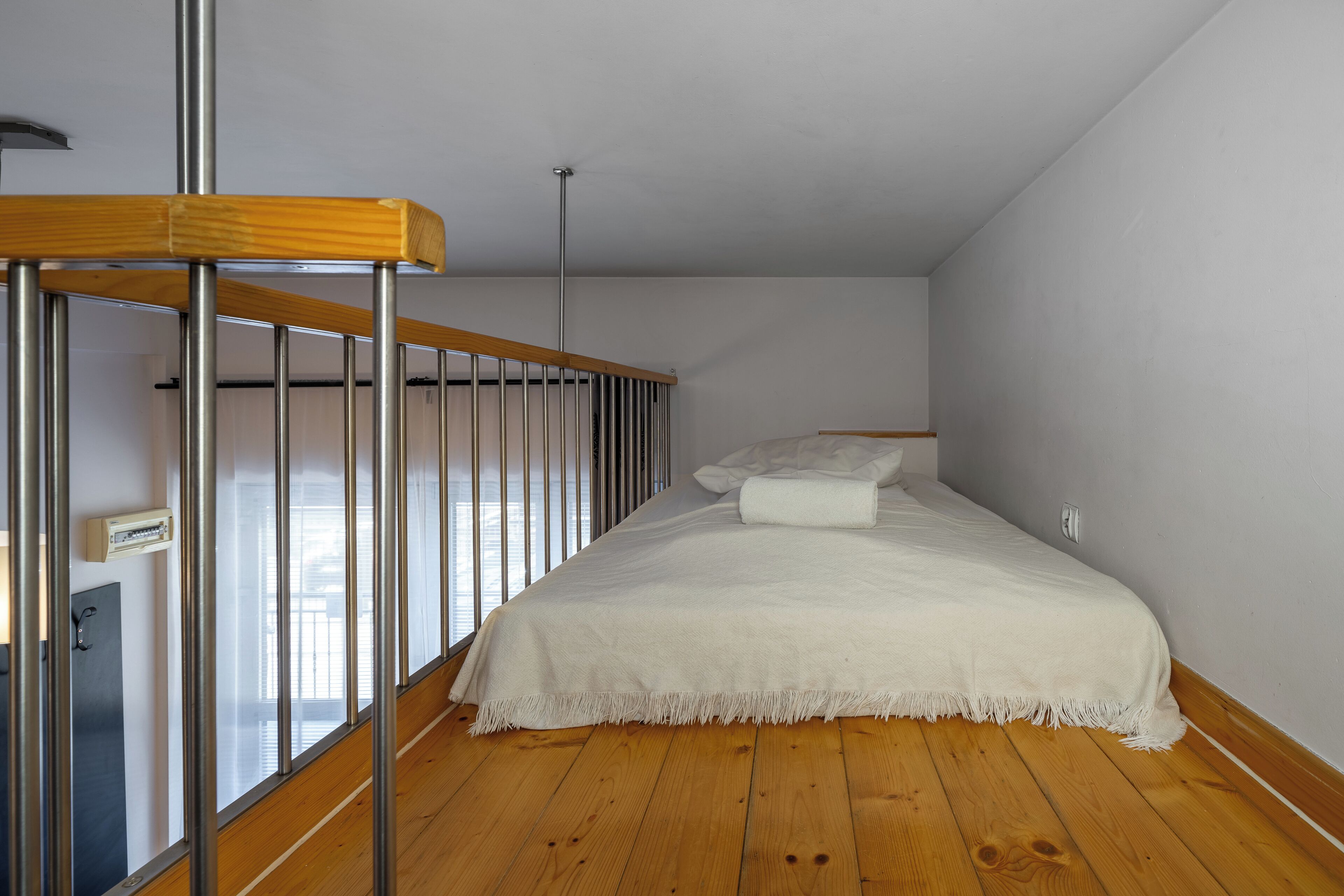 A bedroom with a wooden floor, a white bed with a white throw, and a metal railing.