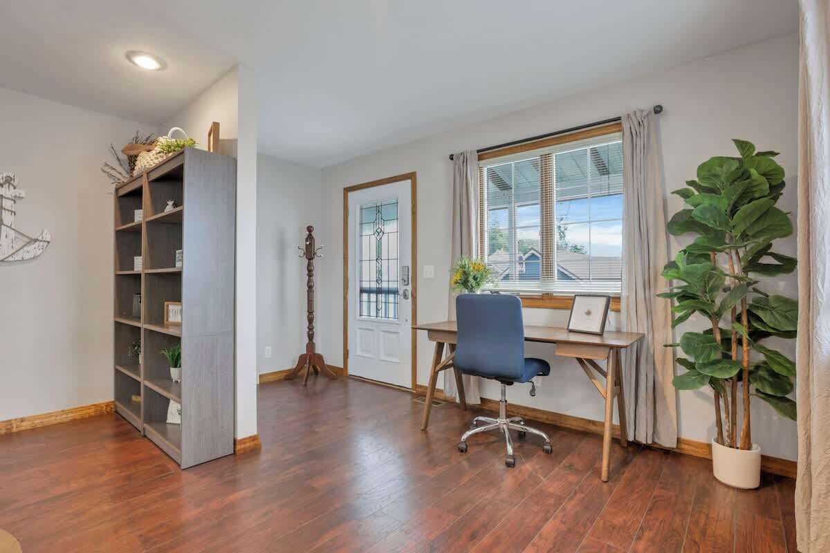 Une pièce avec un plancher en bois, un bureau avec une chaise, une bibliothèque, une plante en pot et une fenêtre avec des rideaux.
