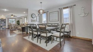 Un vaste salon comprenant un coin repas, avec une grande table en bois et des chaises assorties, un ventilateur de plafond et une vue sur l’extérieur.