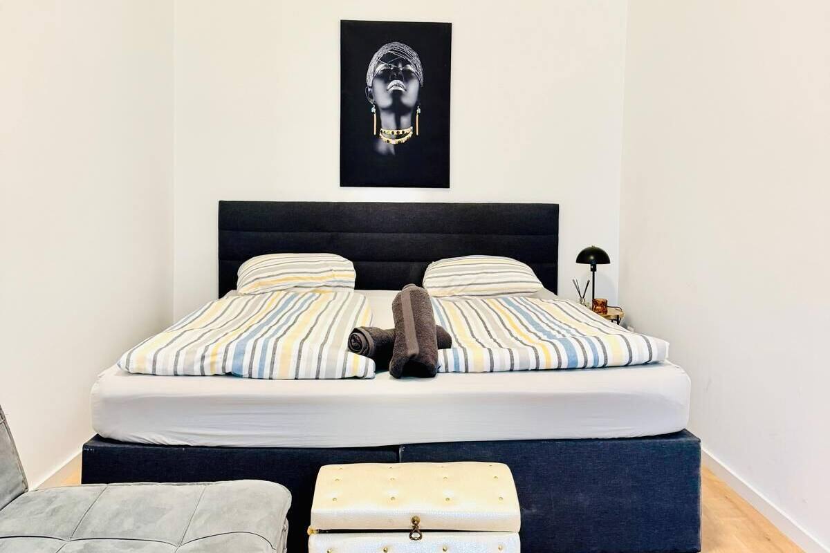 Een slaapkamer met een bed, nachtkastje, lamp, grijze fauteuil en een dressoir.