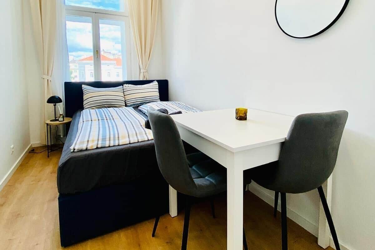 Een kleine kamer met een bed, een tafeltje, twee stoelen, een ronde spiegel en een raam met gordijnen.