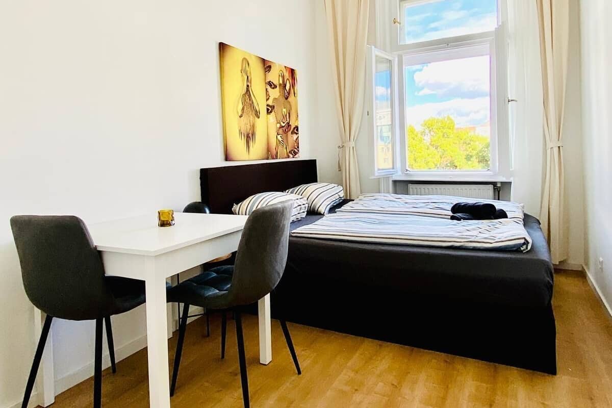 Een slaapkamer met een bed, een bureau en stoelen. Er is een raam met gordijnen, en een schilderij aan de muur.