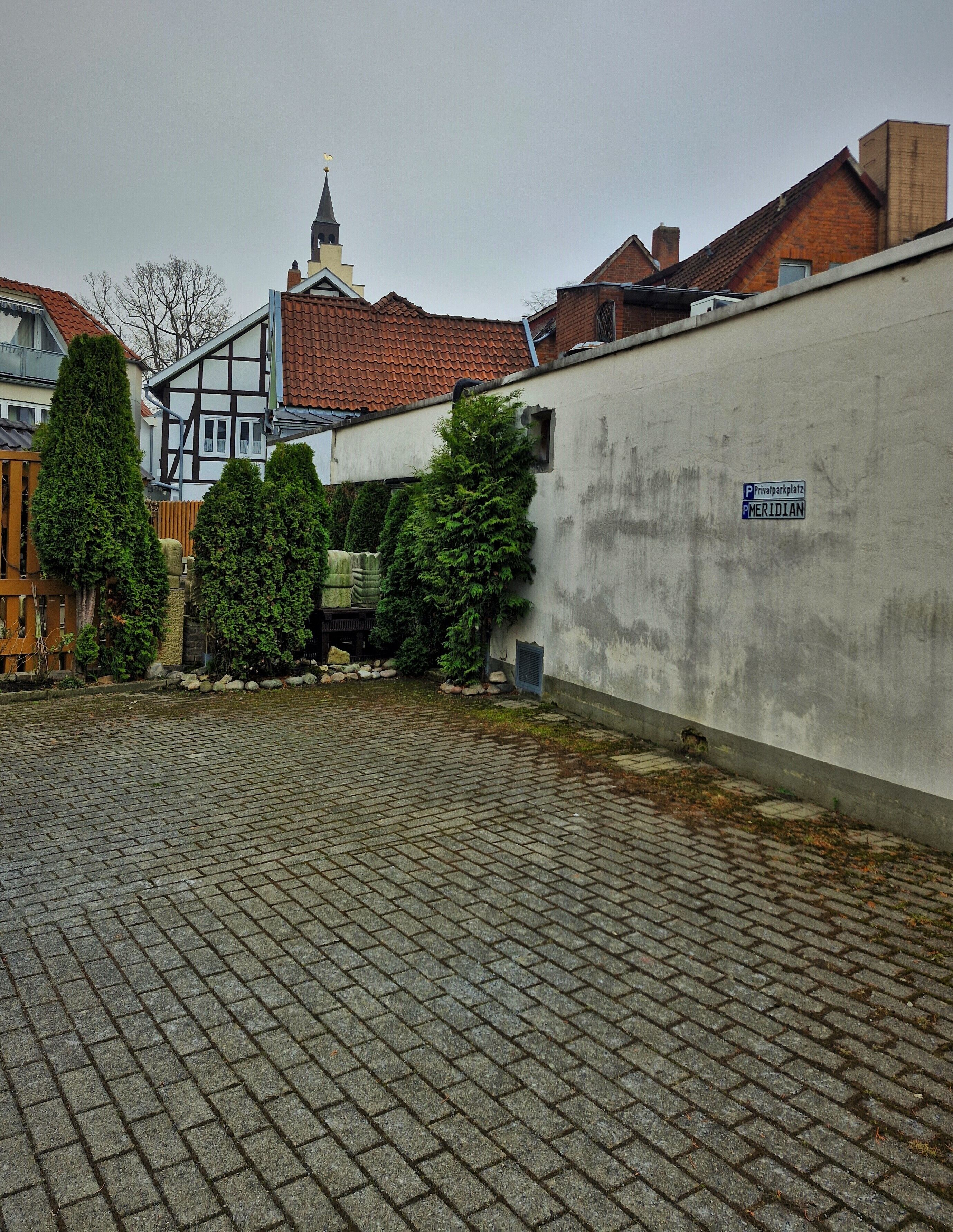 Un cortile pavimentato con un muro bianco, un cartello con la scritta "Privatparkplatz MERIDIAN" e il campanile di una chiesa sullo sfondo.