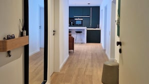 A modern hallway with a wooden floor, a large mirror, and a kitchen area in the background.